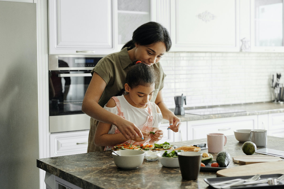A woman stands behind her daughter at a kitchen counter. They are preparing a meal together.